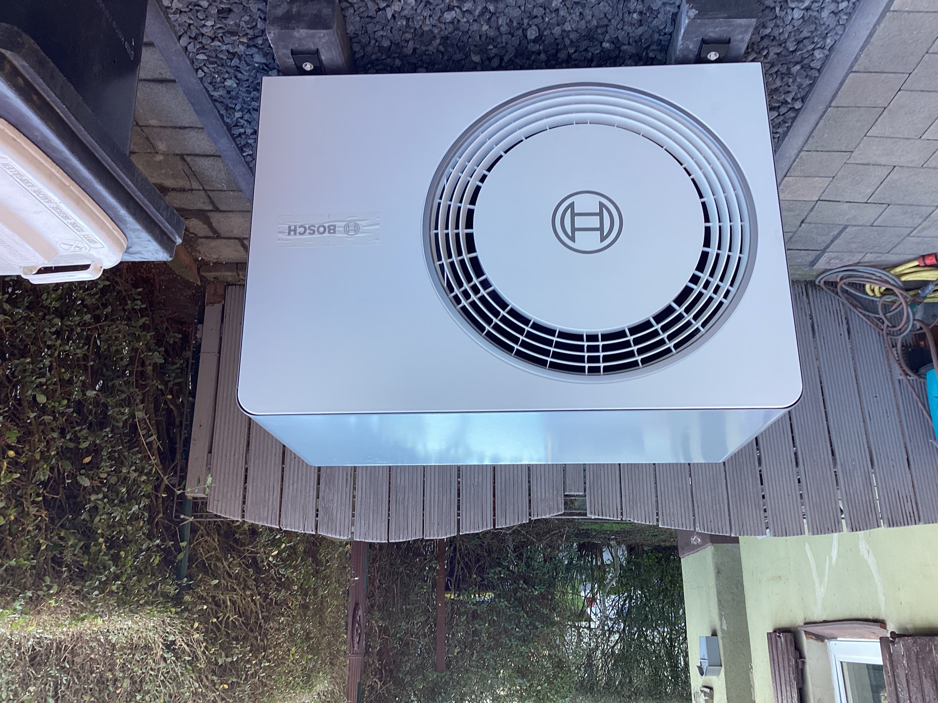 Eine weiße Bosch-Wärmepumpe steht auf einem Kiesbett neben einer Holzbank und ist von Hecken und Sichtschutz umgeben.